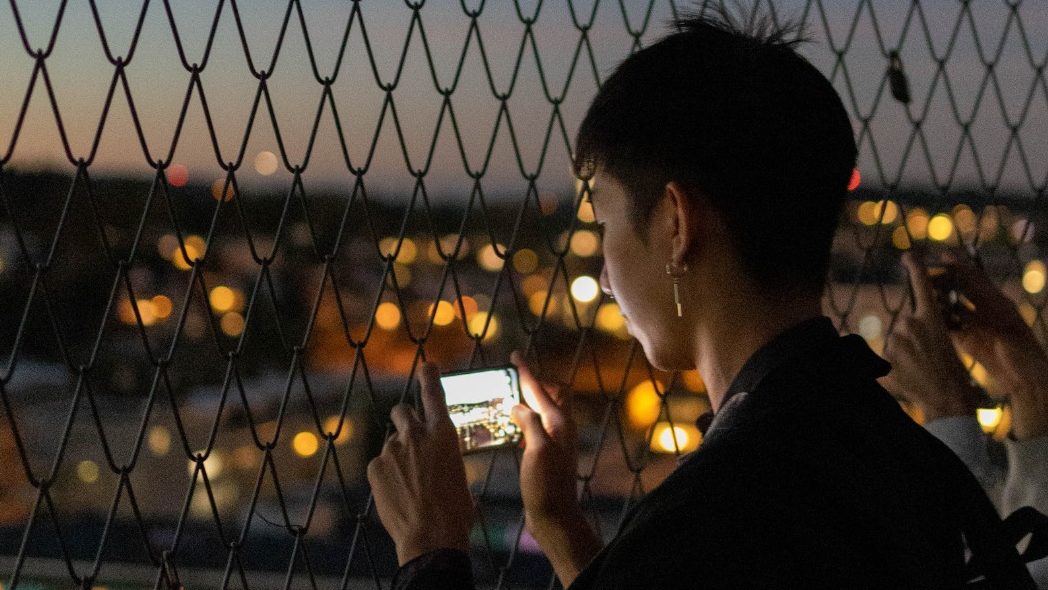 A side view of Michael Tan taking a scenic night picture of Oerlikon with his phone behind a fence and blurred twinkling city lights in the background