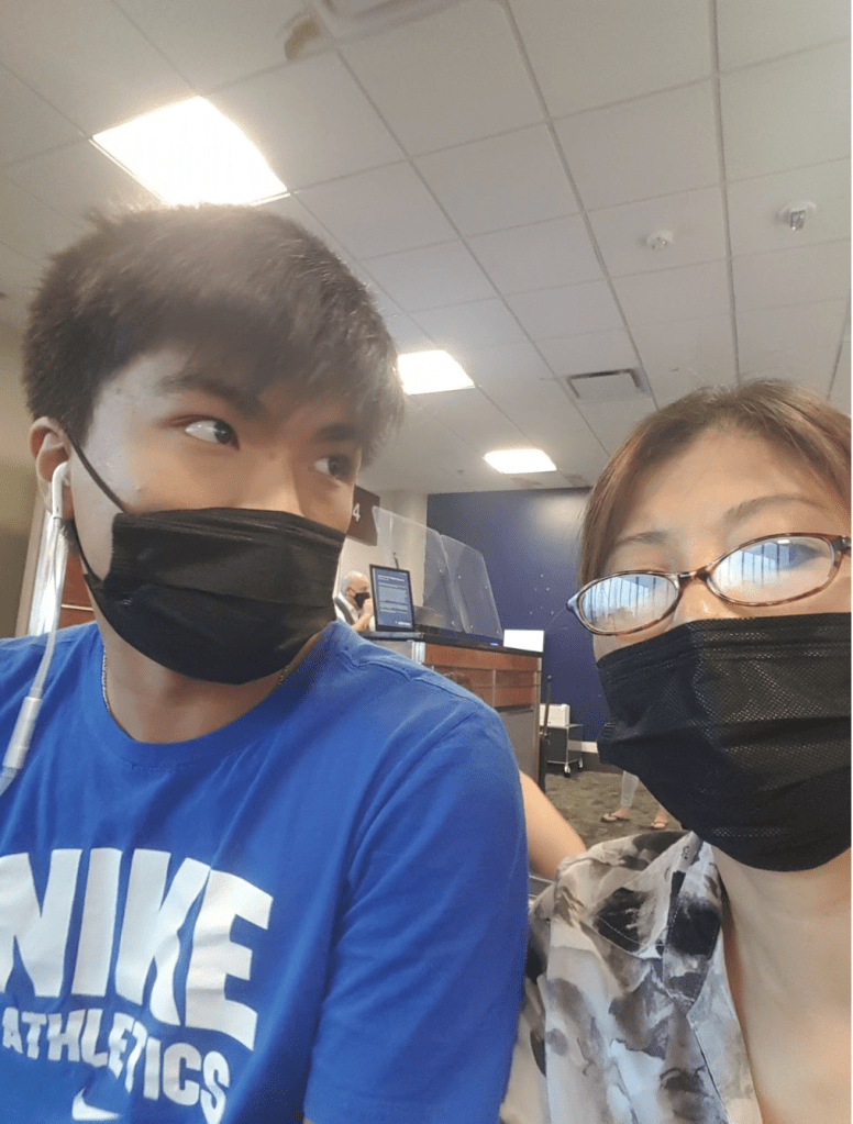 Franklin Nguyen and his mum in black masks. Franklin looks away from the camera to the right and both waiting to board a flight with warm ceiling lights above them and a blue background
