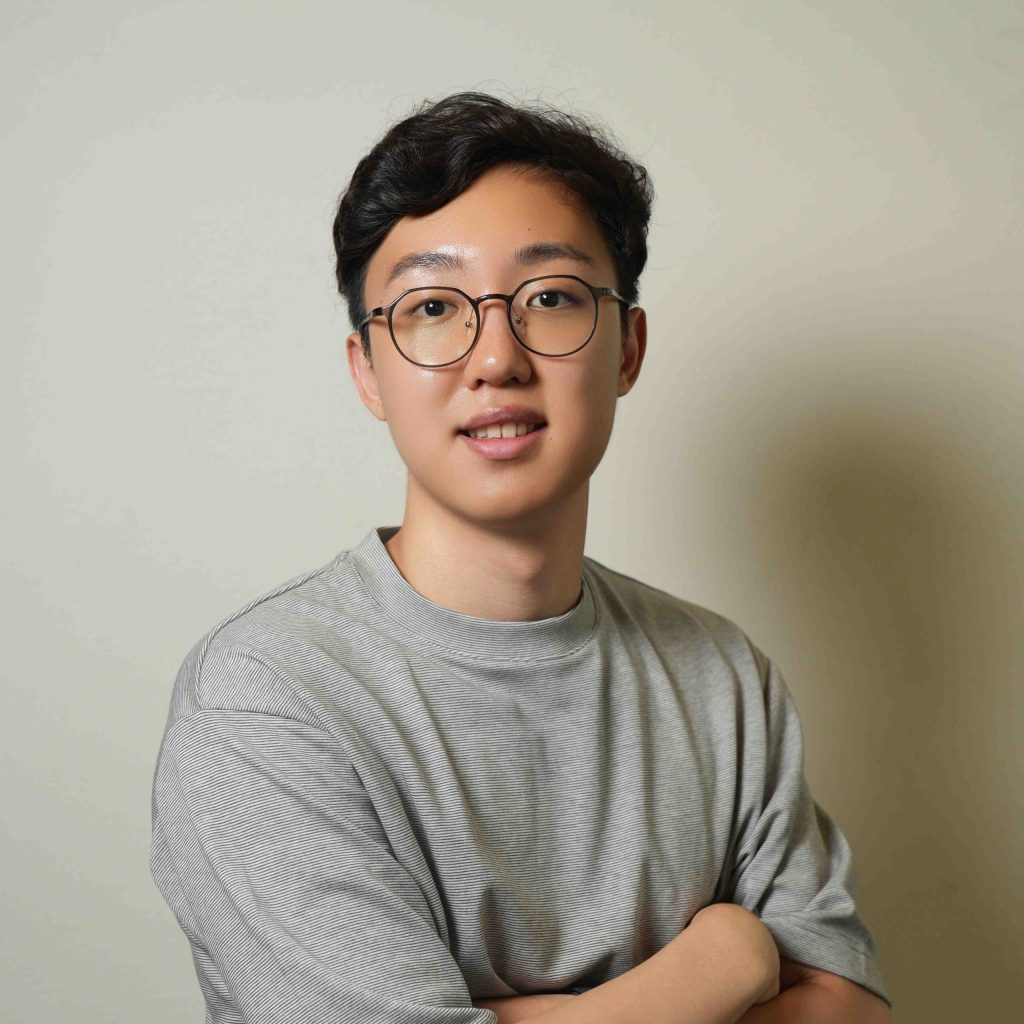 Headshot of Juni C.K. in a grey shirt with black glasses and arms folded